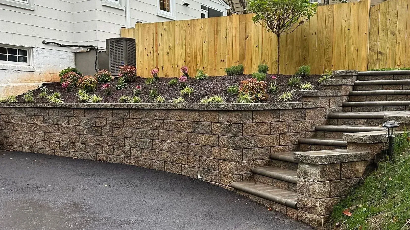 Stone retaining wall with flower bed and wooden fence beside outdoor steps on a sloped driveway Stone retaining wall with flower bed and wooden fence beside outdoor steps on a sloped driveway