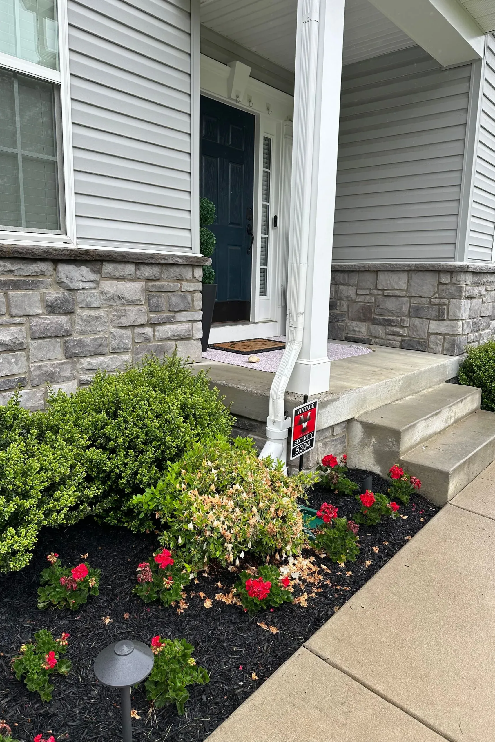 Front entrance of a house with stone facade, steps, and colorful flower beds along the walkway