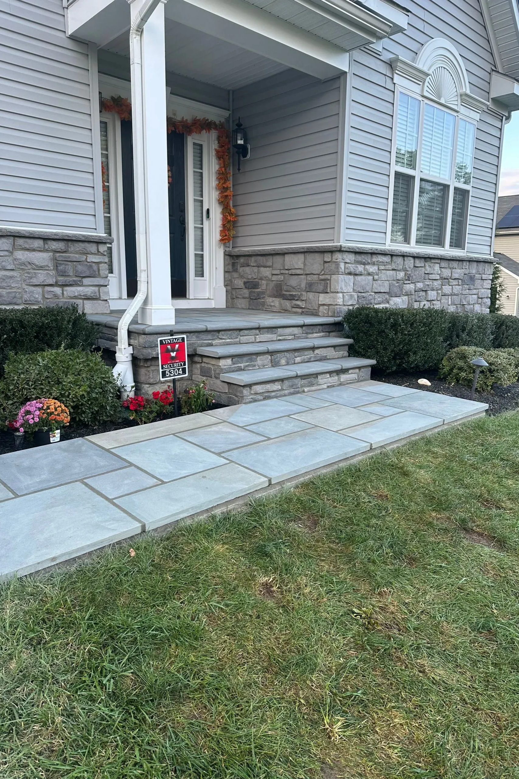 Front entry of a house with stone steps, walkway, shrubs, and a small red yard sign.