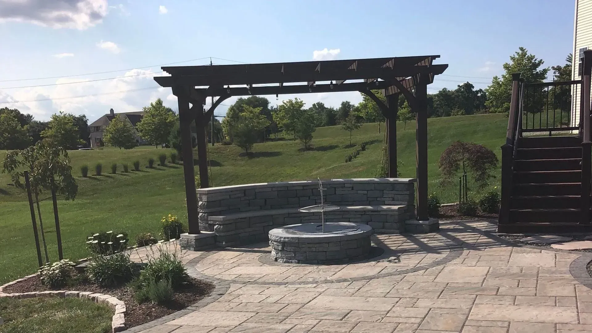 Stone patio with circular fire pit and wooden pergola overlooking a grassy landscape Stone patio with circular fire pit and wooden pergola overlooking a grassy landscape