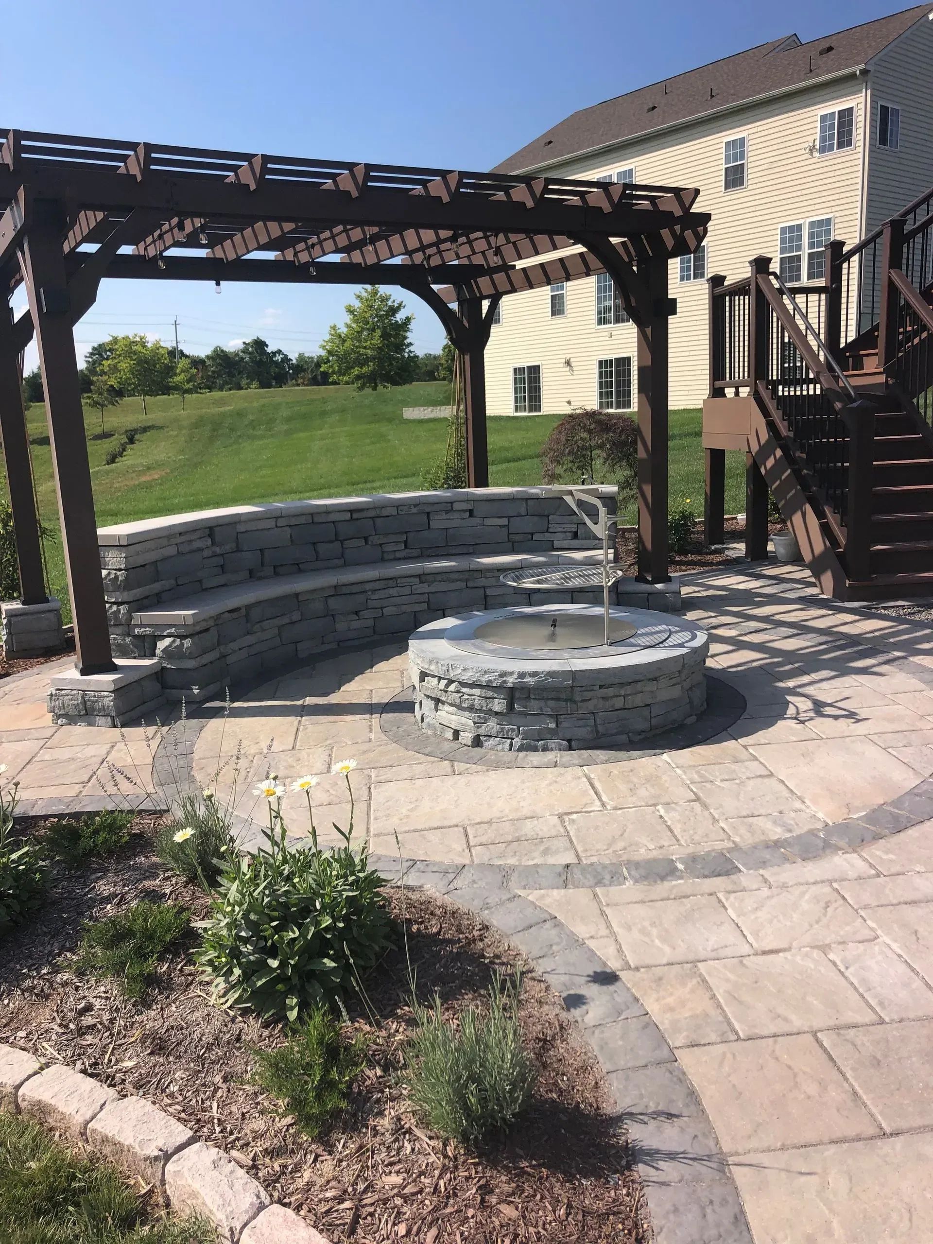 Stone fire pit and curved seating under a wooden pergola in a backyard patio beside a house.