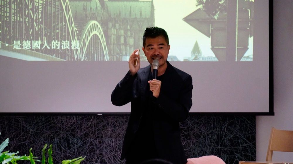 身穿黑色夾克的男子對著麥克風講話，同時指向投影影像。