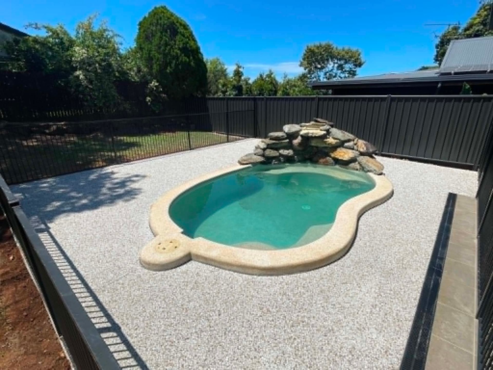 Small backyard pool with turquoise water, rock waterfall feature, and black fence under a sunny blue sky