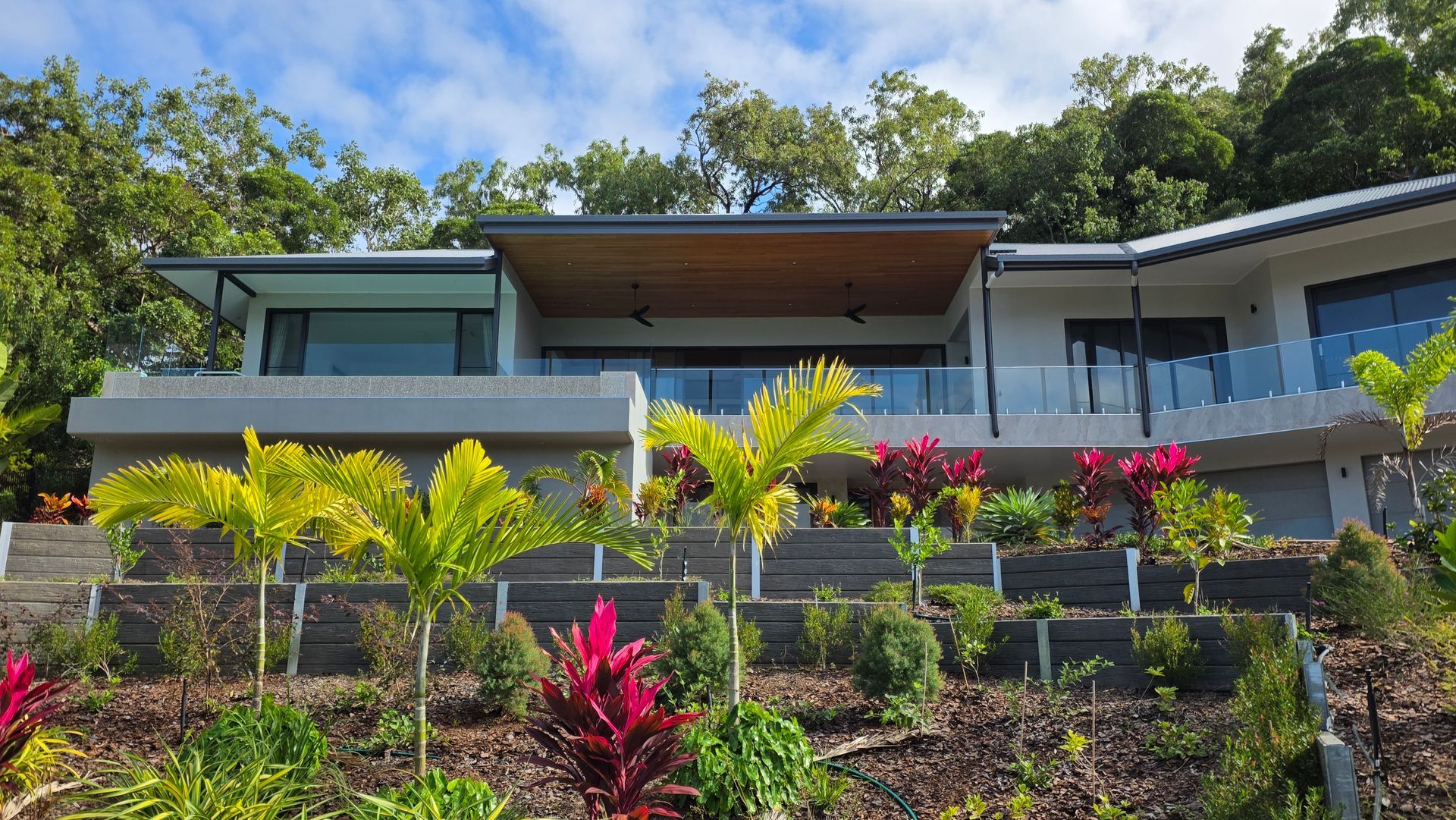 Modern house with large glass balcony, tropical plants, and colorful landscaping under a bright sky