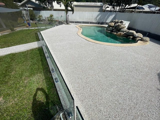 Backyard pool with curved rock waterfall and large white concrete patio beside green lawn