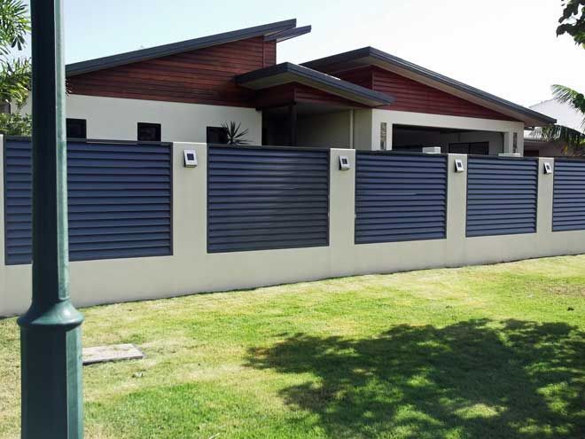 Modern house with red roof behind a white wall and blue gates, seen from a grassy street.