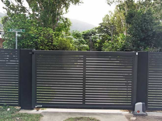 Gray metal driveway gate with horizontal slats, flanked by dark pillars and greenery behind it