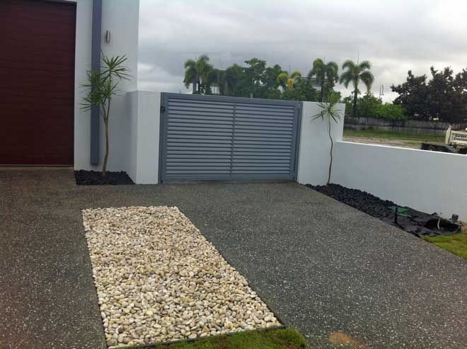 Driveway entrance with gray gate, stone path, and gravel yard beside a white wall and palm trees.