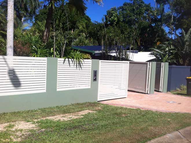 Modern house entrance with white louvered fence, sliding gate, and driveway beside palm trees.