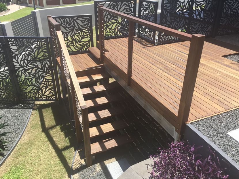 Wooden ramp with railing and stairs beside a deck, bordered by decorative black metal panels and gravel.
