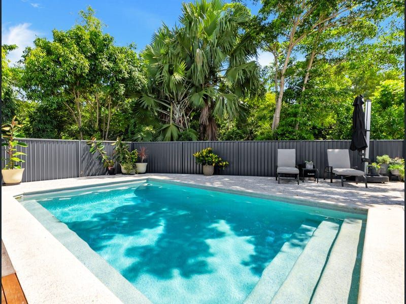 Sunny backyard pool with turquoise water, gray deck chairs, and lush green trees behind a dark fence