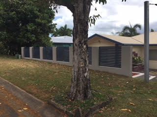 Residential house with a beige fence and louvered panels, viewed from the street with a large tree in front.