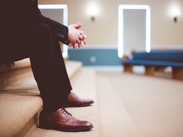 Person seated on church pew, hands clasped, in a softly lit room with blue walls and windows