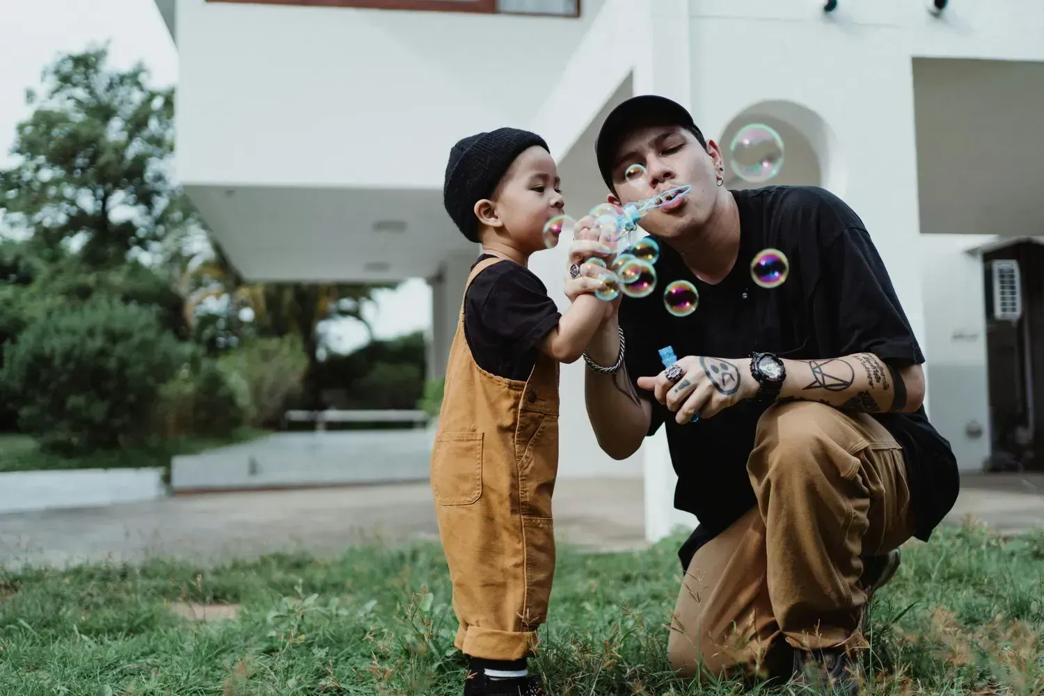 Child and adult kneeling on grass, blowing bubbles outside a modern house