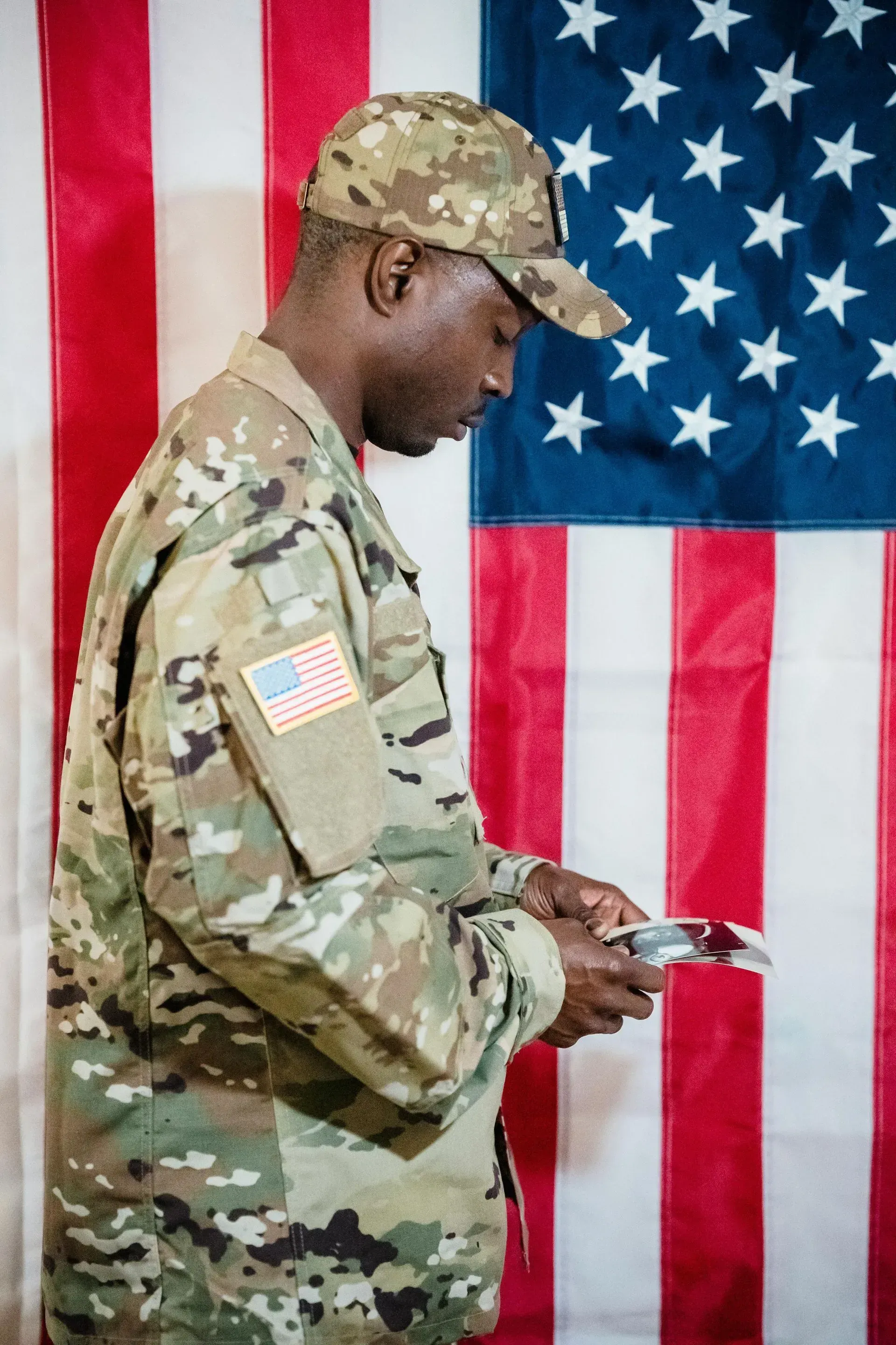Soldier in camouflage standing in profile before a U.S. flag, holding a small object
