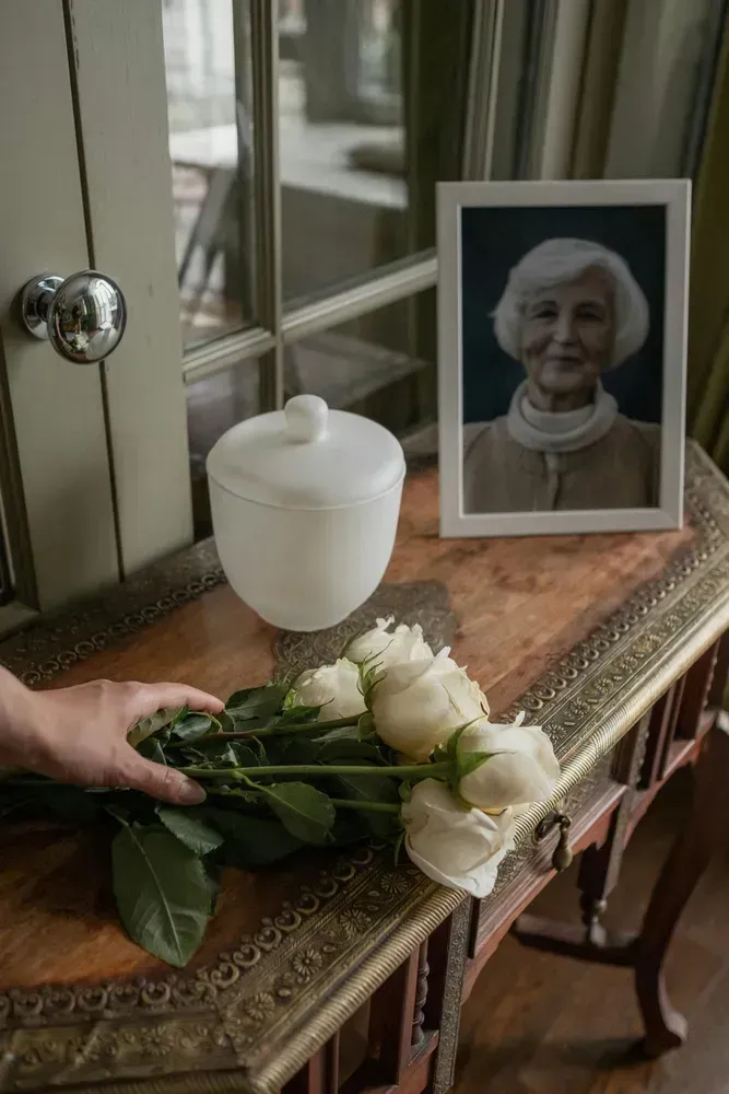 White roses and a framed portrait on a table beside a window, with a hand placing flowers nearby