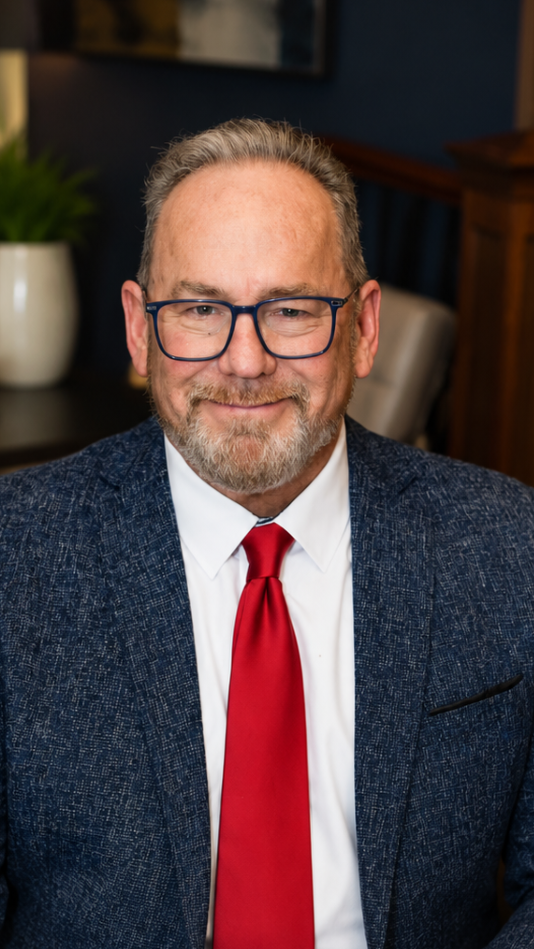 Portrait of a smiling man in glasses, navy blazer, white shirt, and red tie against a blurred indoor background
