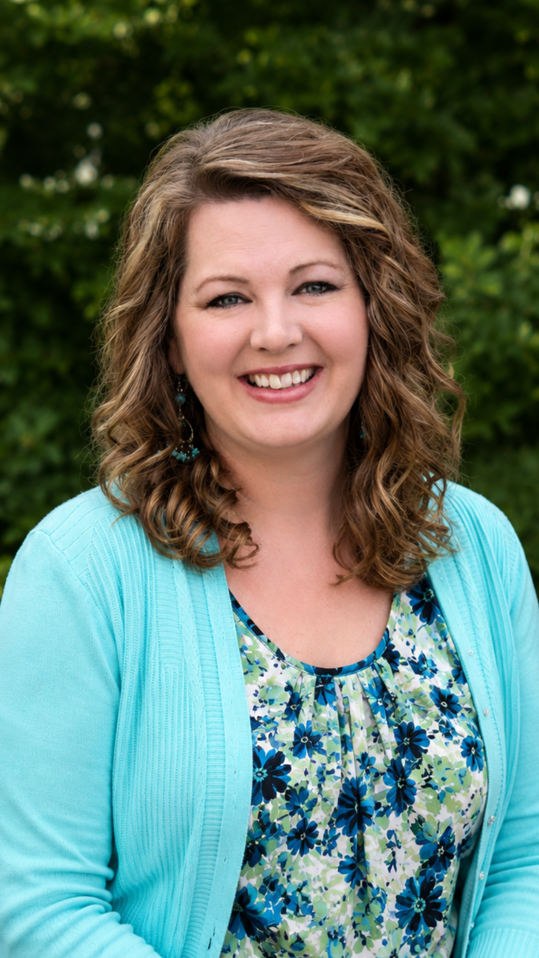 Smiling woman in a turquoise cardigan and floral top, posed outdoors against a green background