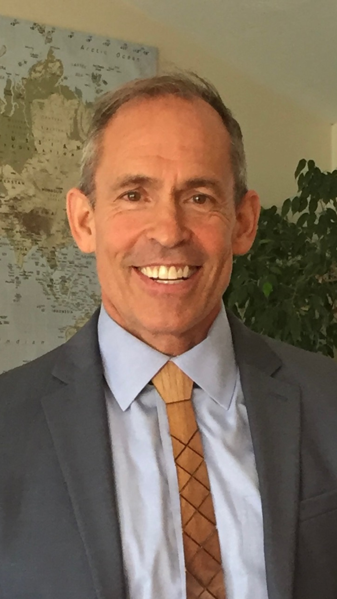 Smiling man in a gray suit and orange tie standing indoors near a wall map and plant