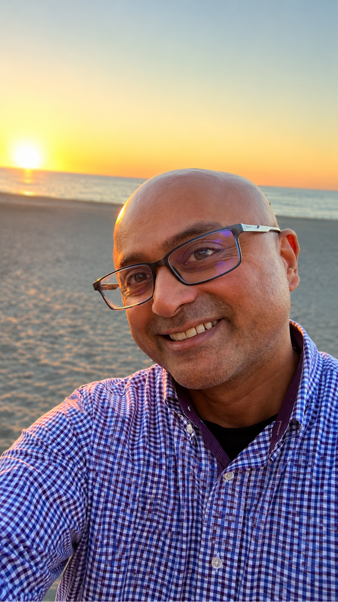 Smiling person in a blue checkered shirt taking a selfie on a beach at sunset