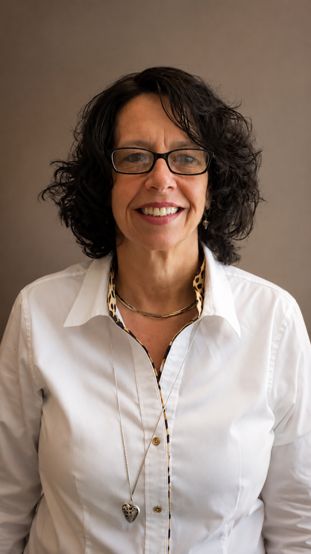 Smiling woman with glasses and dark curly hair wearing a white blouse and necklace on a plain background