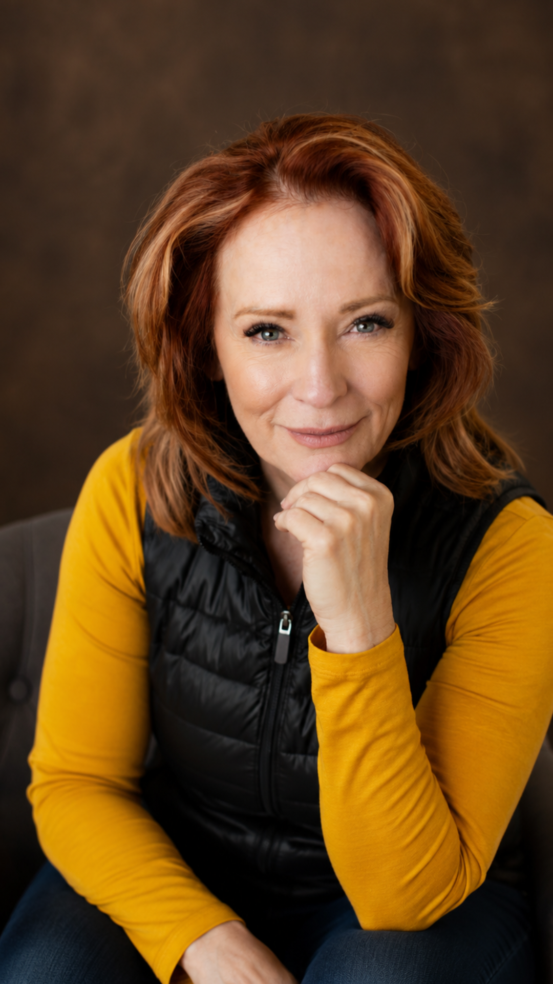 Portrait of a smiling woman in a black vest and mustard sweater, resting her chin on her hand.