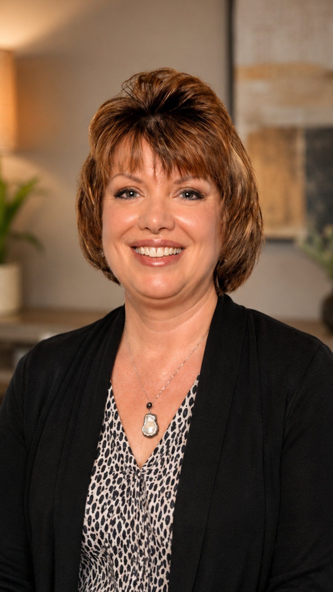 Smiling woman in a black cardigan and patterned blouse, posed indoors against a blurred background.