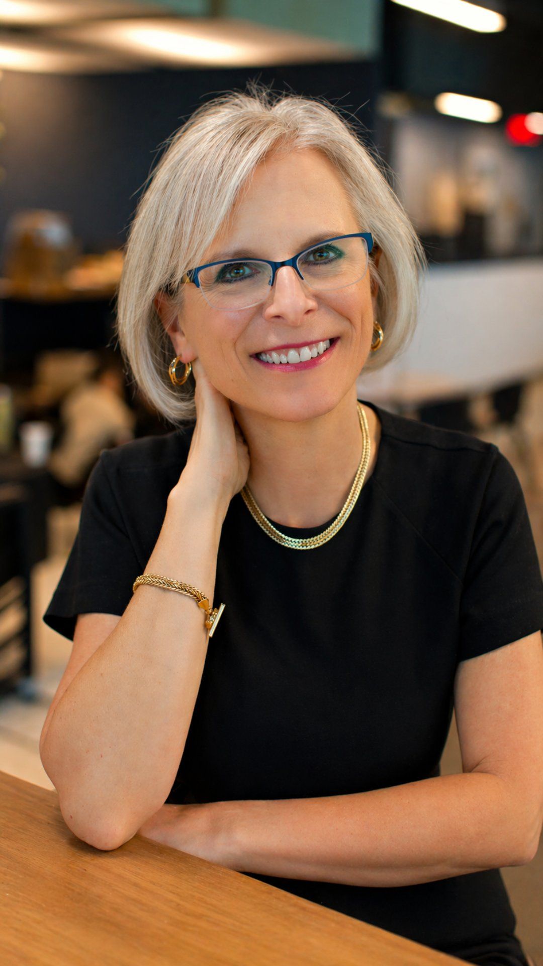 Smiling woman with glasses and short gray hair, seated in a black top in a softly blurred indoor setting