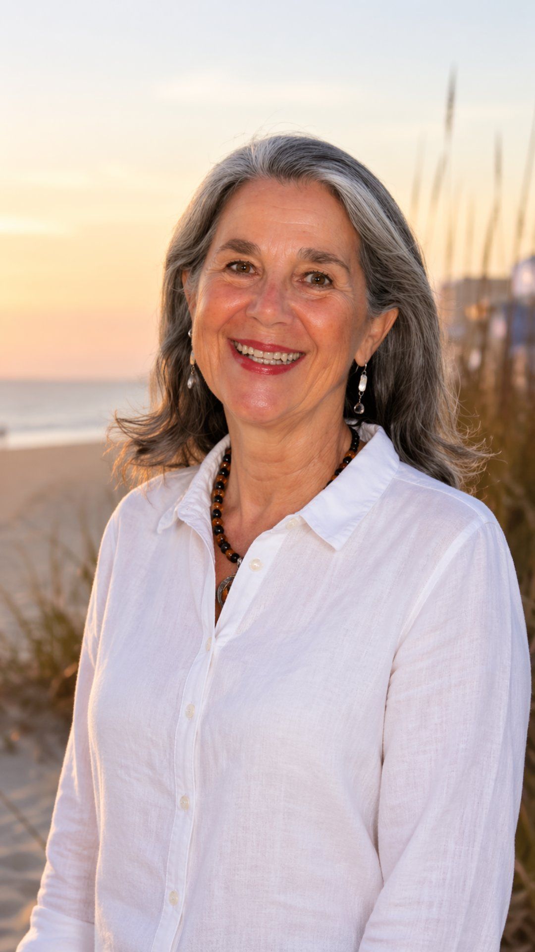 Smiling woman in a white shirt standing on a beach at sunset with tall grasses in the background