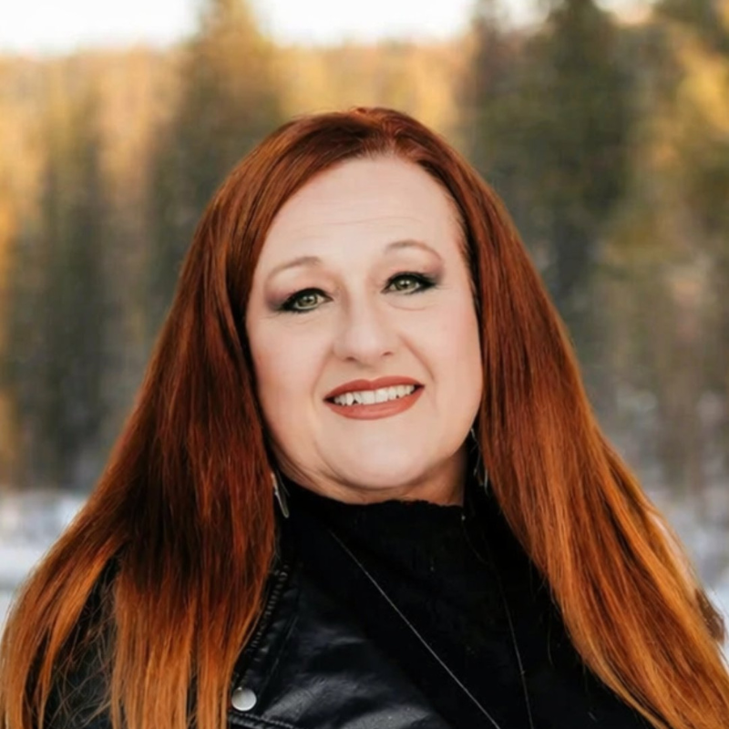 Portrait of a smiling woman with long red hair in a black jacket, standing outdoors against a blurred forest backdrop