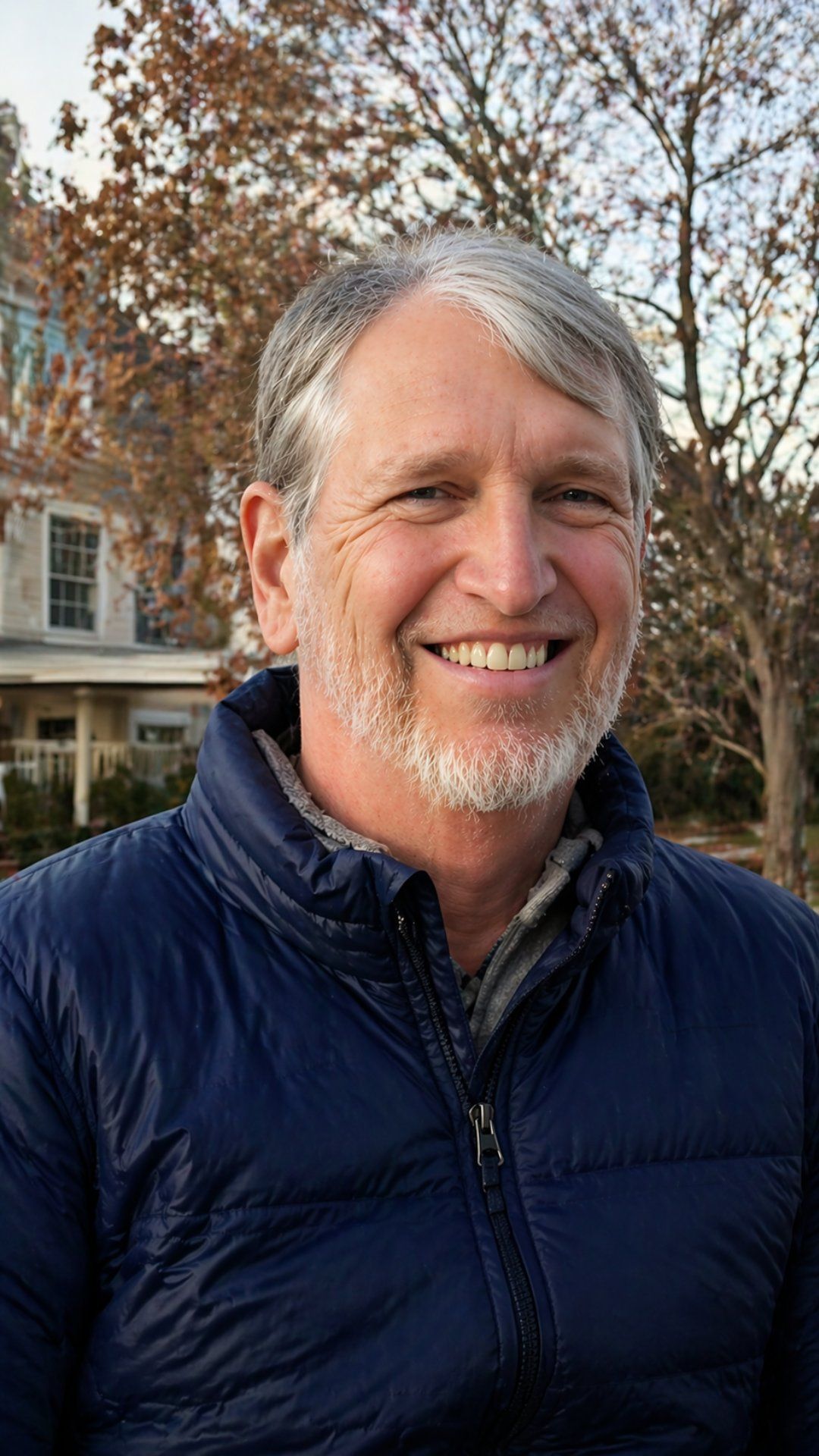 Smiling person in a navy jacket standing outdoors with trees and a house in the background