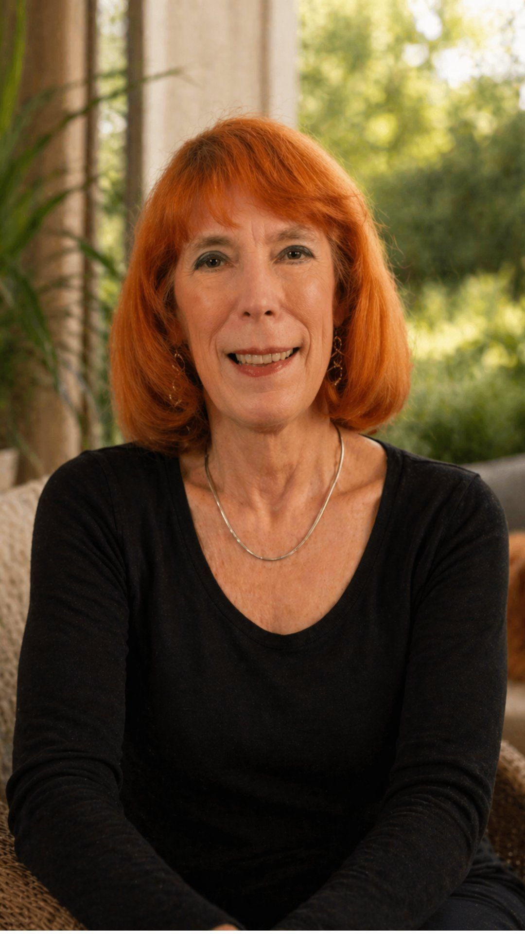 Portrait of a smiling woman with red hair in a black top, seated indoors by a bright window