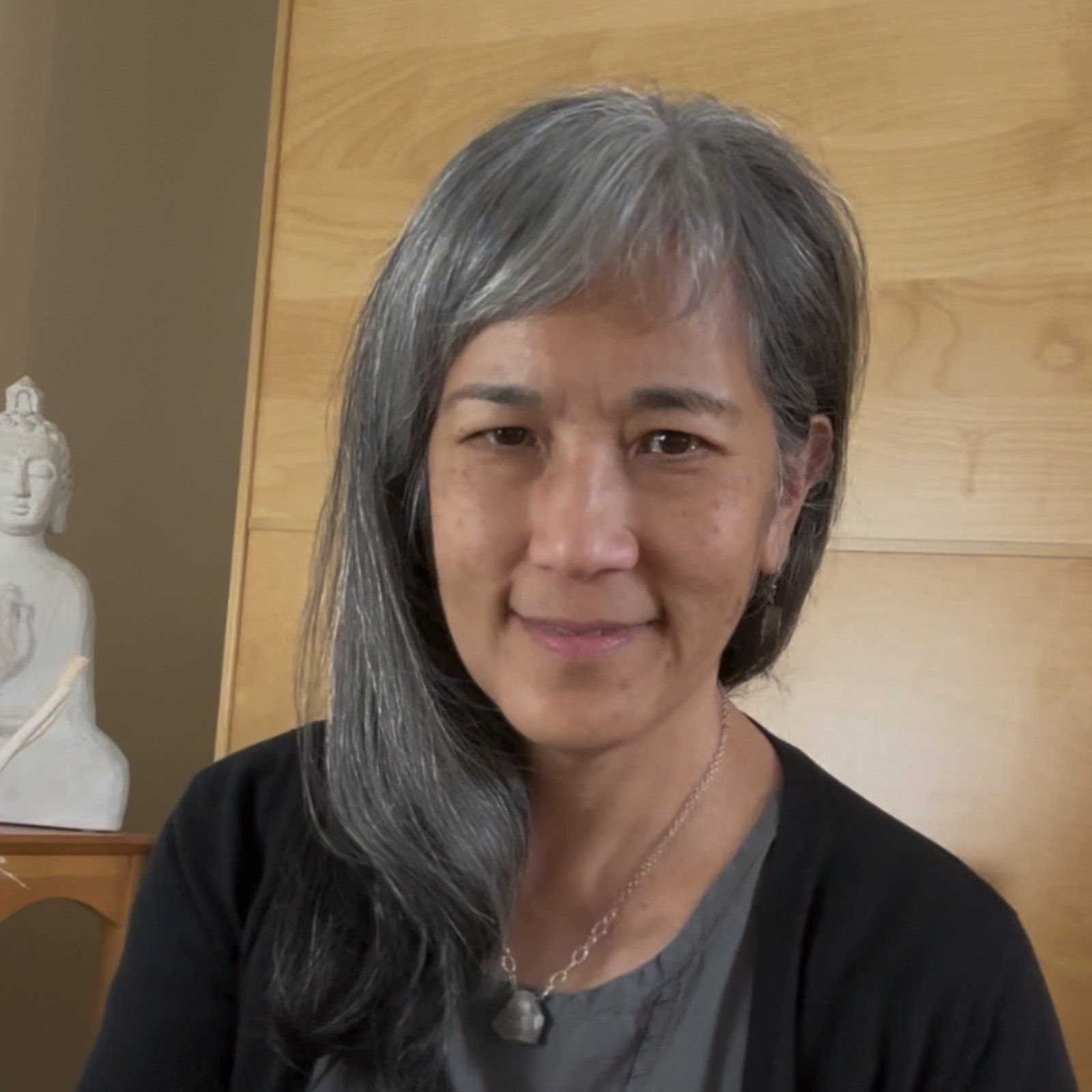 A person with long, grey hair and a calm expression sits indoors, with a small Buddha statue visible in the background.