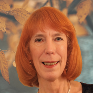 Woman with red hair smiling, small gold earrings, silver necklace. Blurred fall leaves background.
