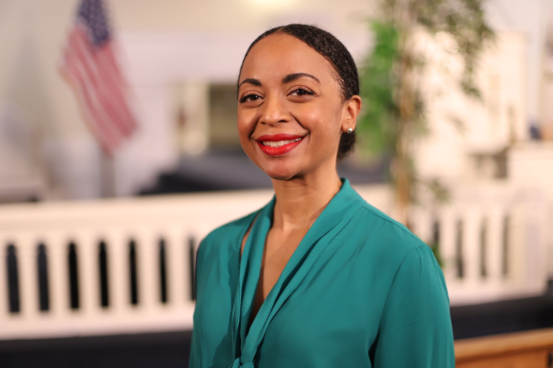 A woman in a green shirt is smiling in front of an american flag.