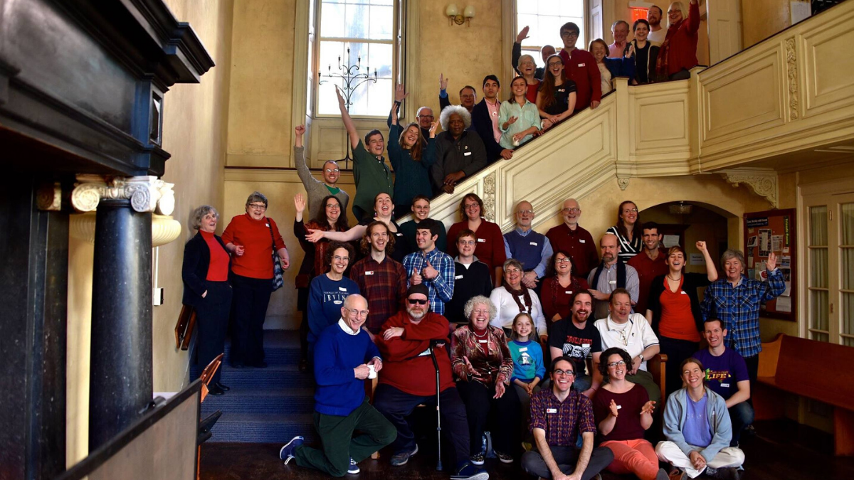 A large, cheerful group poses on and around a wide wooden staircase in a building with high ceilings and warm lighting.