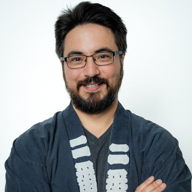 A person with glasses and a beard smiling while wearing a dark blue, patterned kimono-style garment against a white wall.