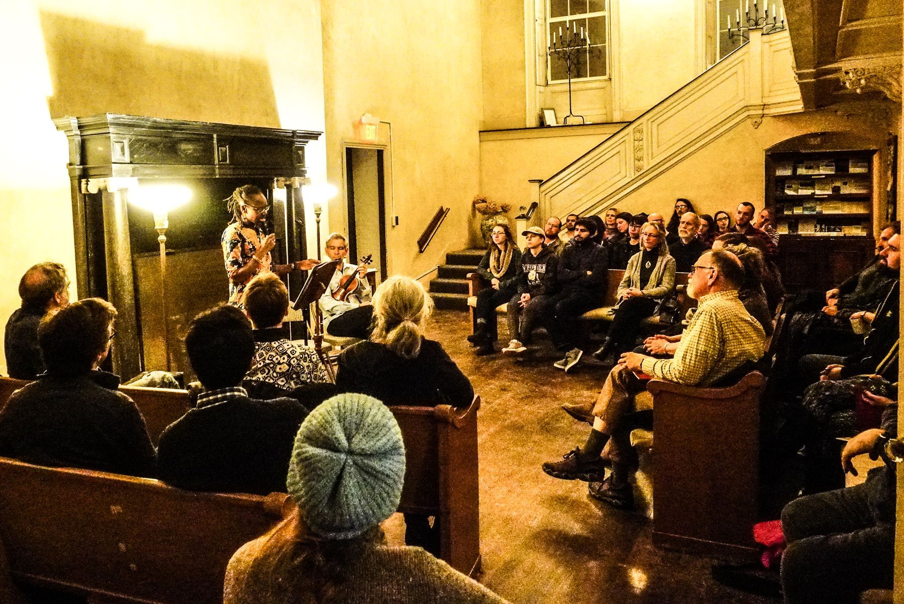 A performer plays guitar for a seated audience in a dimly lit, high-ceilinged room with a large fireplace and staircase.