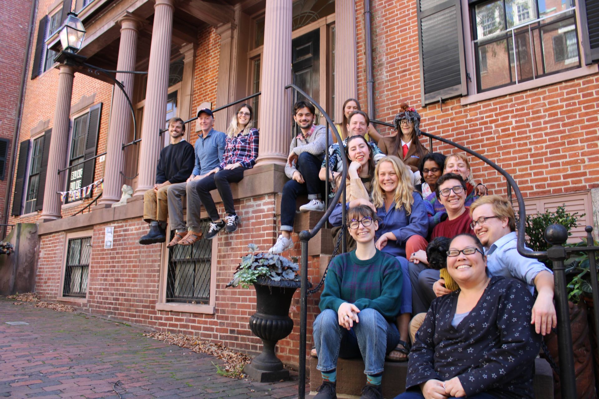 Fifteen people pose on the front steps and columned porch of a red brick building on a bright day.