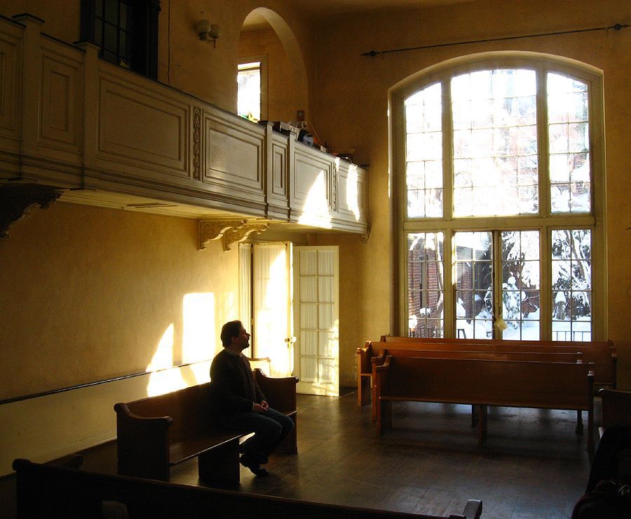 Man sits in a sunlit church. Wooden pews, balcony, tall arched window.