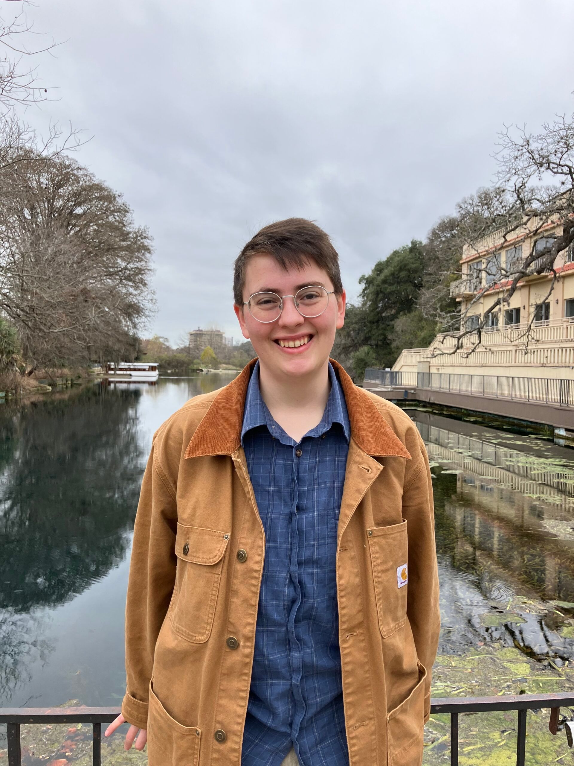 A person wearing glasses and a tan jacket smiles while standing in front of a river and a building.