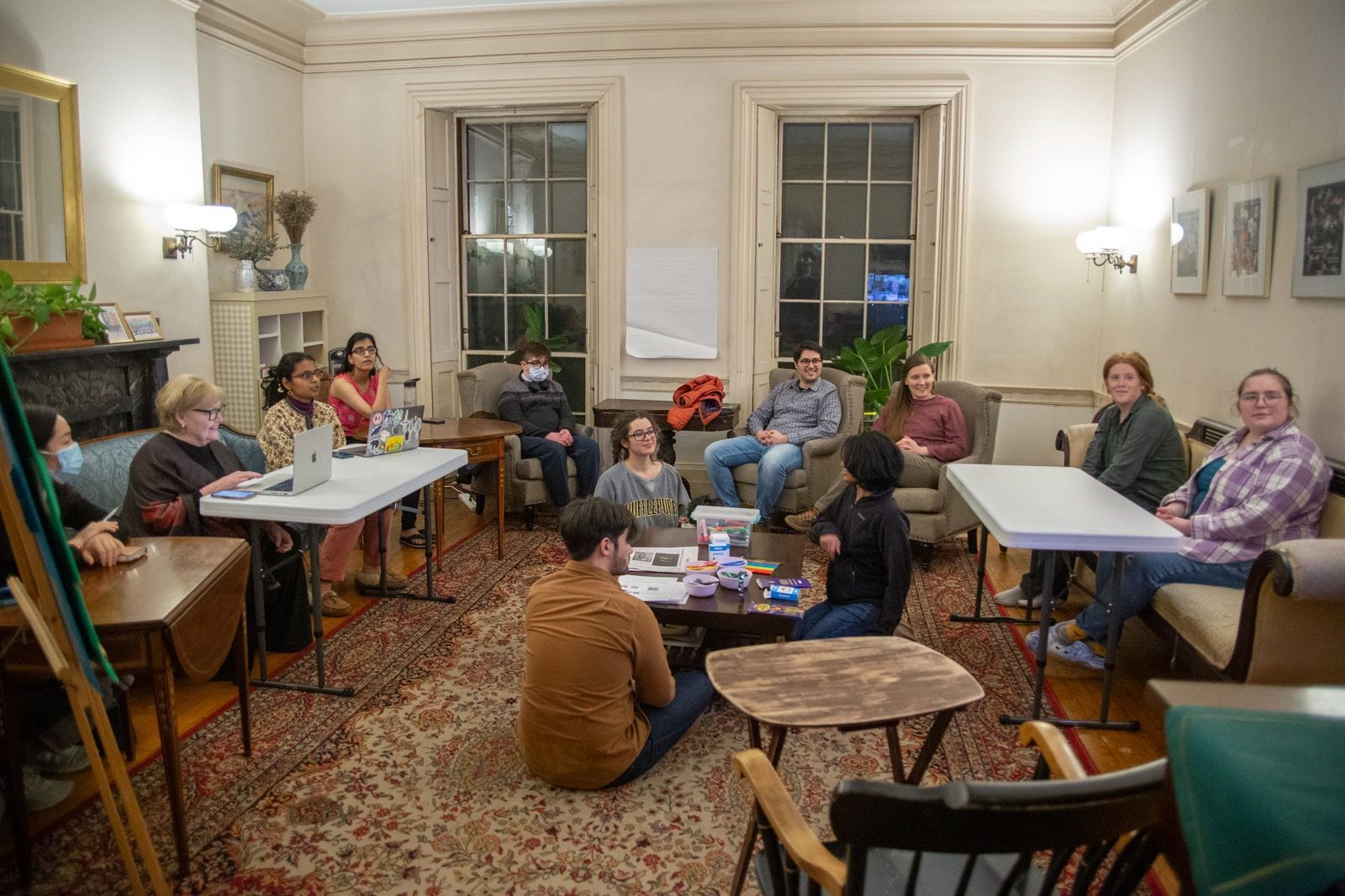 A diverse group of people gather in a well-lit room with patterned rugs for an informal meeting around tables and chairs.