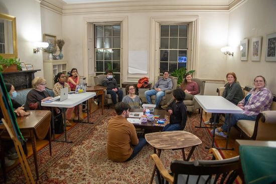 A group of people sitting in a living room, some at tables and some on the floor, during a discussion.
