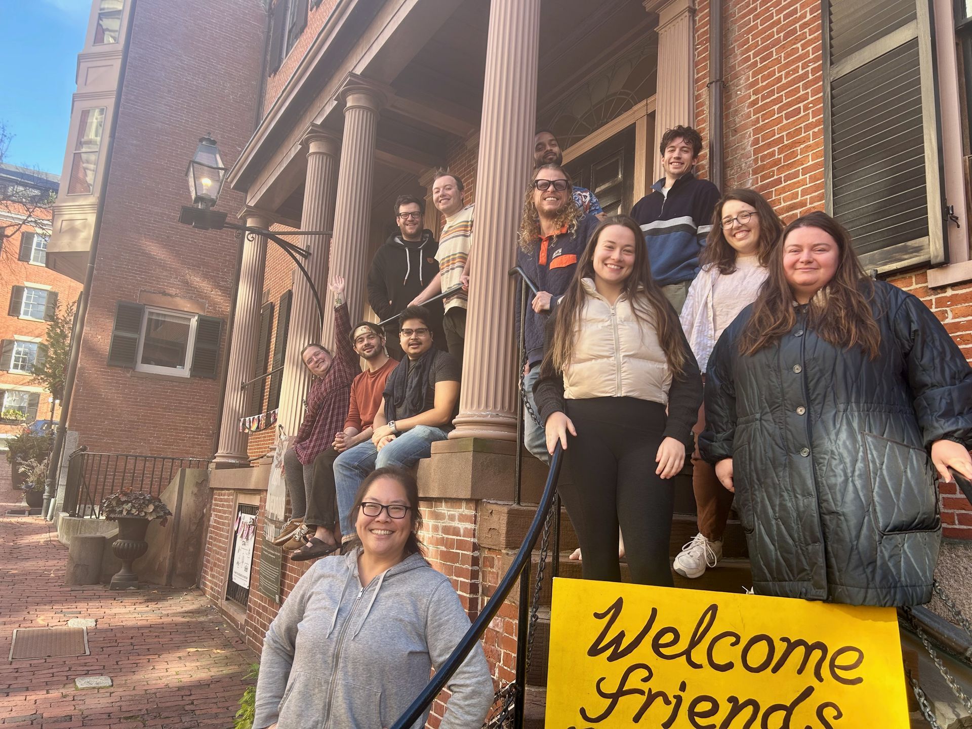 A group of people standing on a brick building's porch stairs behind a yellow sign that reads