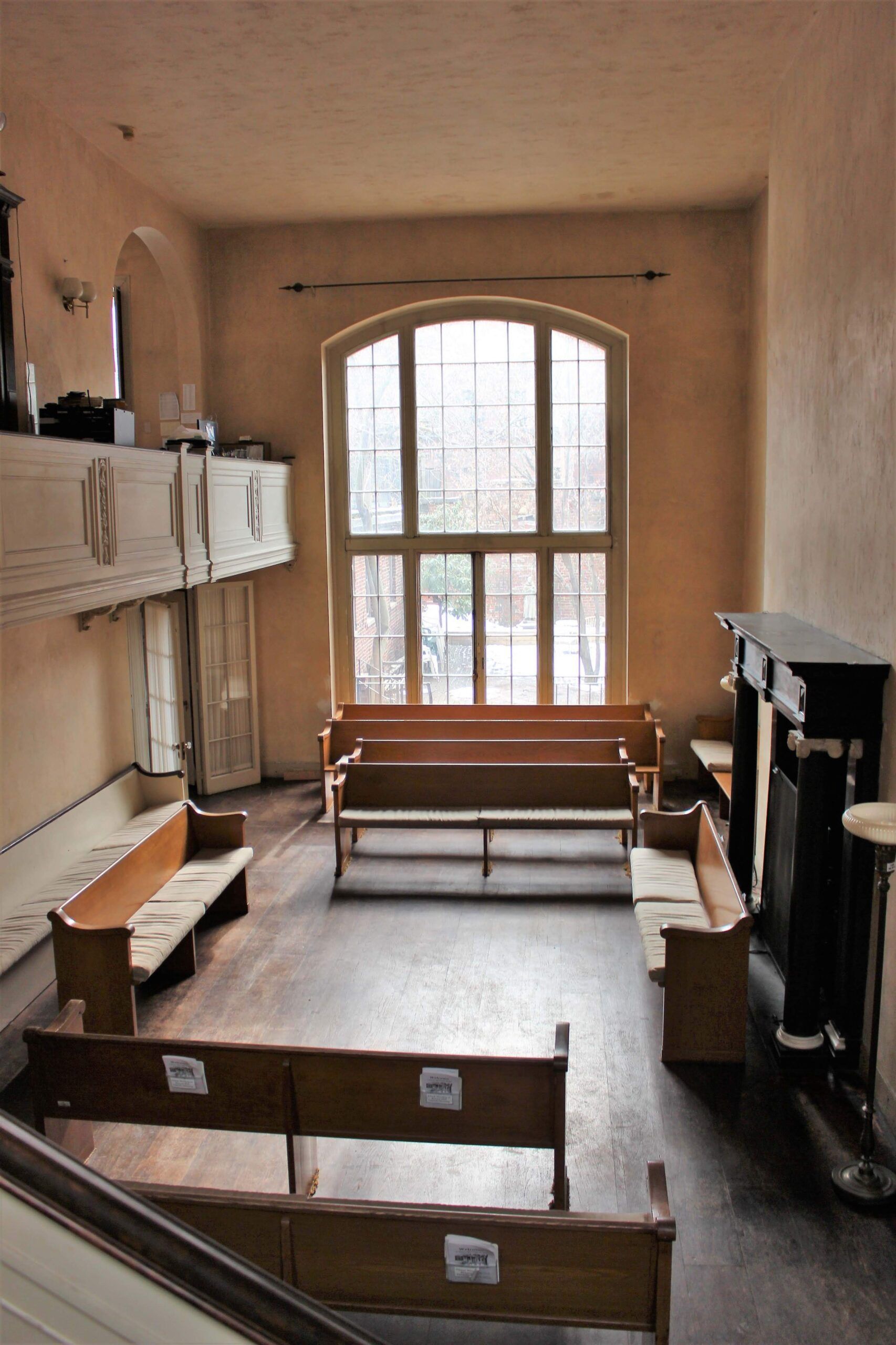 Interior view of a church with wooden benches, a large arched window, and a balcony.
