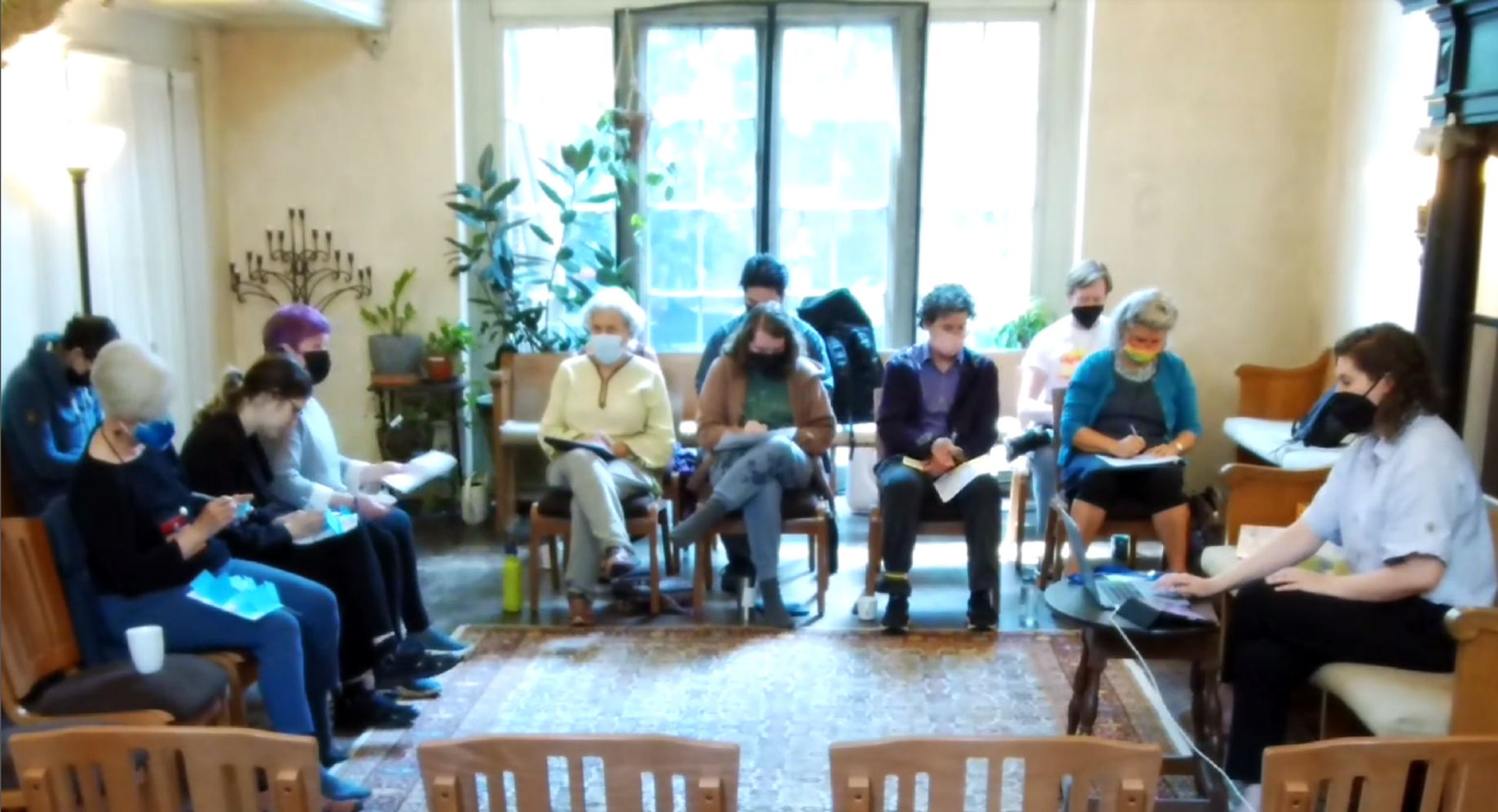 A group of people sit in a circle in a sunlit room, writing on pads of paper during a meeting.