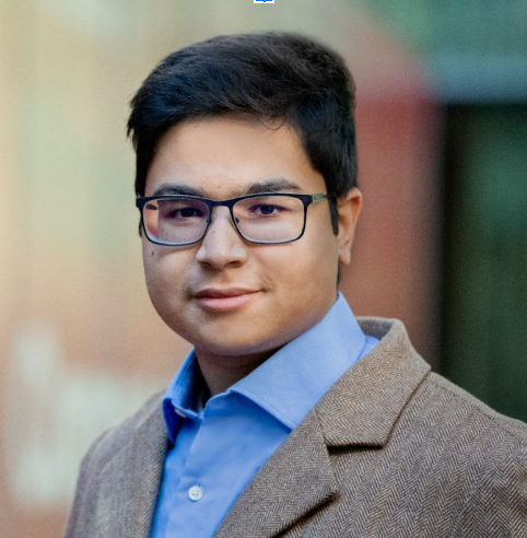A person with short dark hair and glasses wears a blue shirt and a brown blazer, posing against a blurred background.