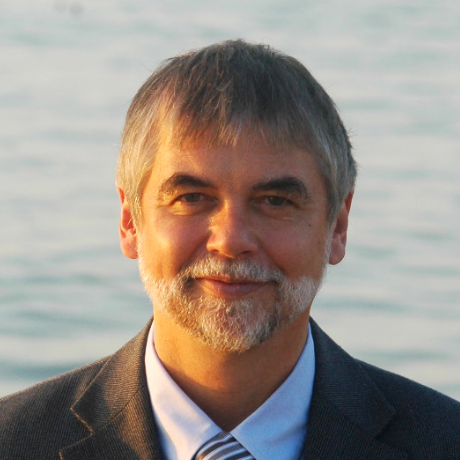 A man with a short beard wearing a blazer and striped tie, smiling gently against a blurred, light-colored water background.