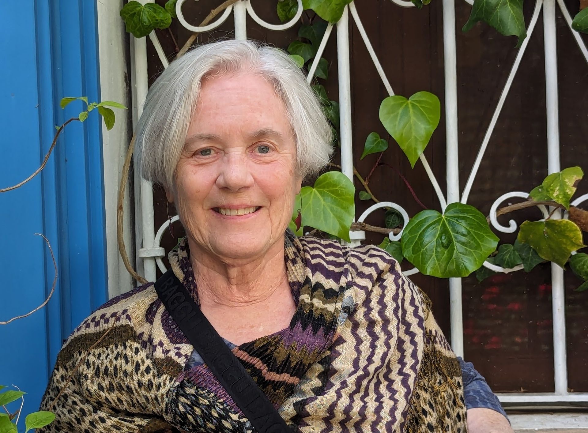 A person with short grey hair smiles in front of a white metal gate covered with green climbing ivy and a blue wall.