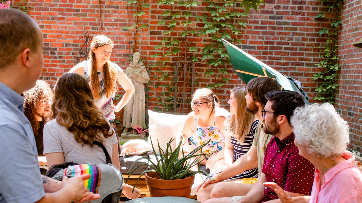 A group of people sits in a circle in a sunlit brick courtyard, facing a woman standing and speaking to them.